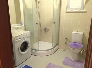 A compact bathroom with a washing machine, a glass-enclosed shower, and a toilet with a purple mat. - Murat Kaptan Apart Otel (Marmara)