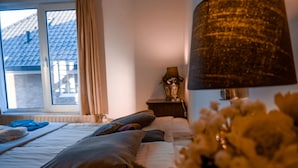 A bedroom with a bed, pillows, a bedside table with a lamp, a window with curtains, and a view of a building outside. - Pension Villa Tanahlot (Zandvoort)