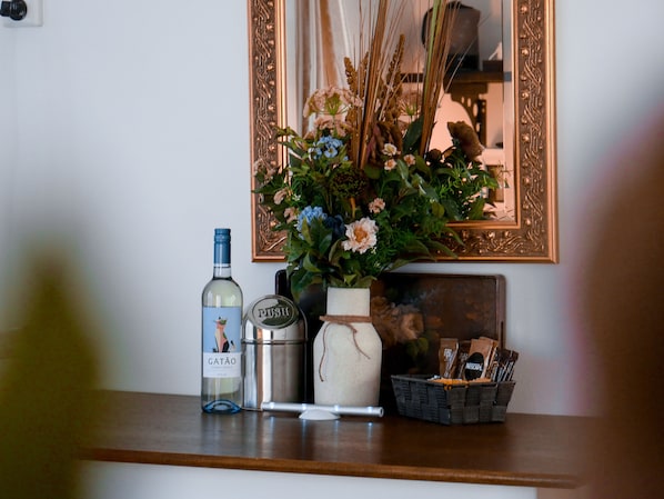 Un estante de madera con una botella de GATÃO, un recipiente metálico, un jarrón pequeño con flores y un cesto de cápsulas de café.