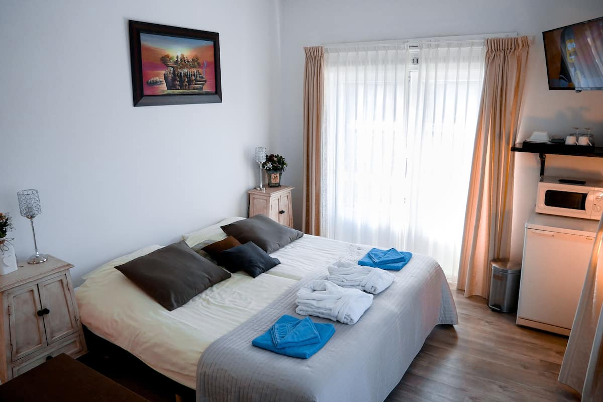 A neatly made bed with pillows, a bedside table with a lamp and flowers, a TV mounted on the wall, and a window with curtains.