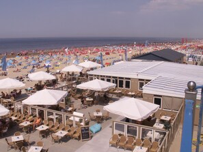 A beachside cafe with outdoor seating and umbrellas, overlooking a crowded beach.