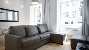 A modern living room with a grey sectional sofa, a small table, and a large window offering a view of buildings outside. - City Stays Sé Apartments (Lisbon)