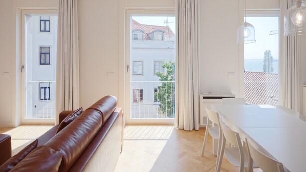 A modern living room with a sofa, a dining table, and chairs. There are large windows with curtains, and a view of buildings outside. - City Stays Sé Apartments (Lisbon)