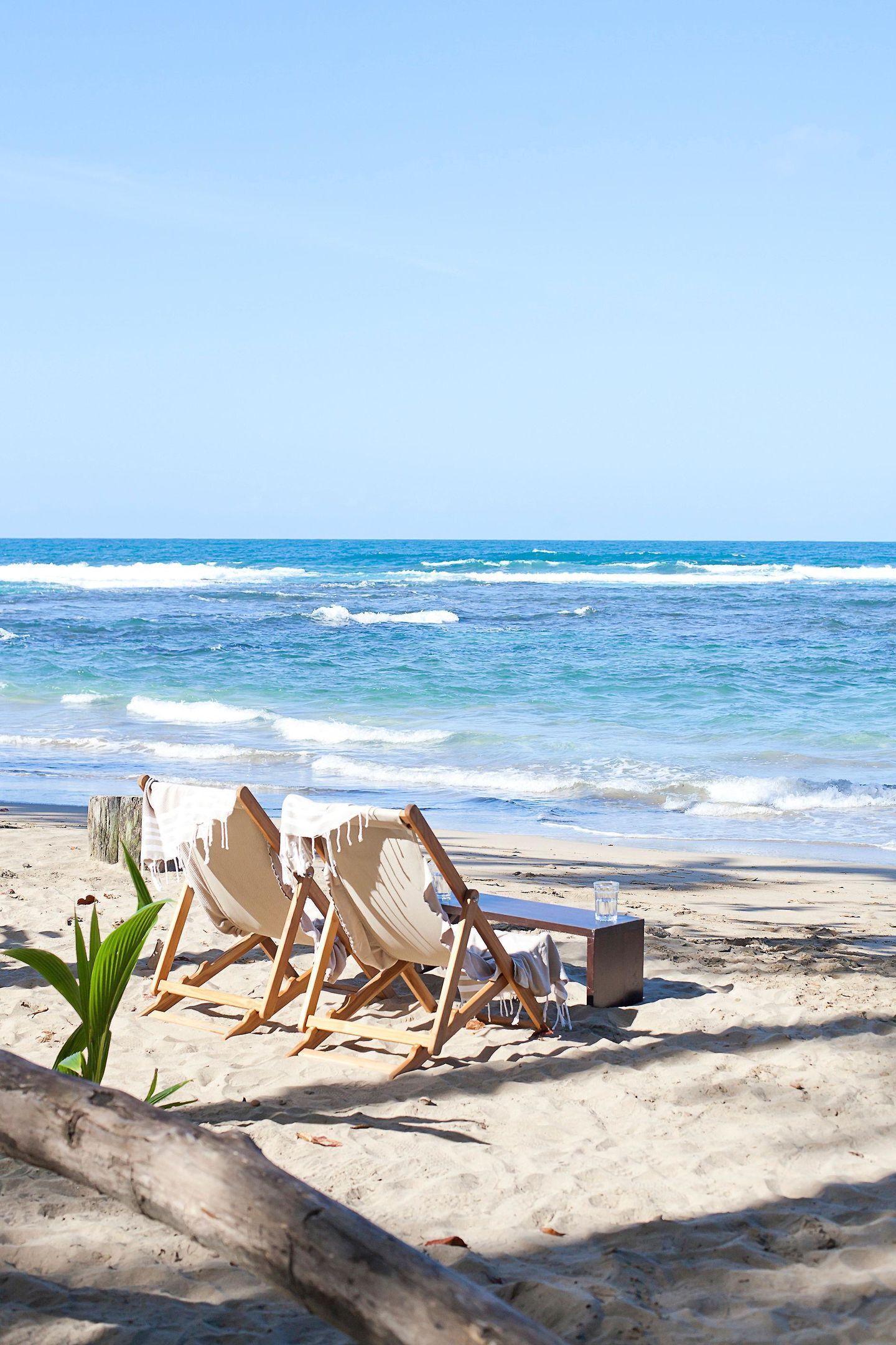 Ein Strand mit Holzliegen, einem Tisch, einer Pflanze und einem Glas Wasser im Sand.
