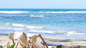 Ein Strand mit Holzliegen, einem Tisch, einer Pflanze und einem Glas Wasser im Sand.