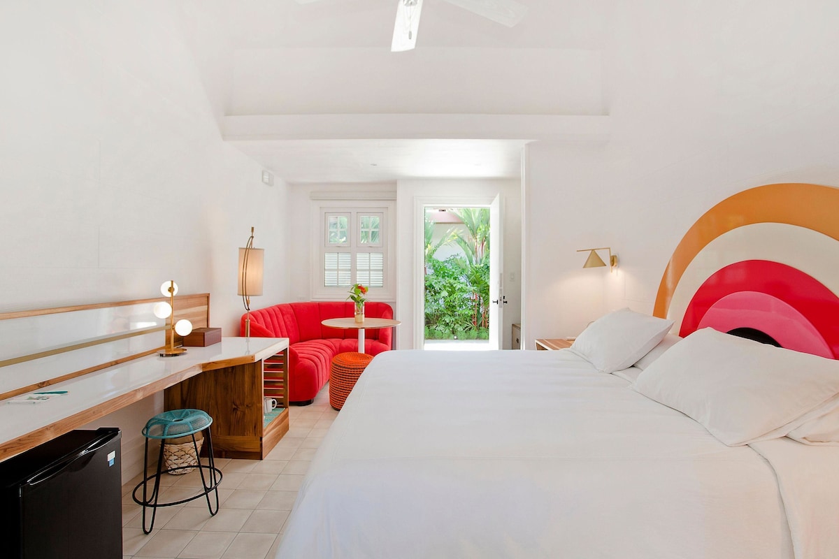 A hotel room with a bed, a desk, a red sofa, a small table, and a view of greenery outside.