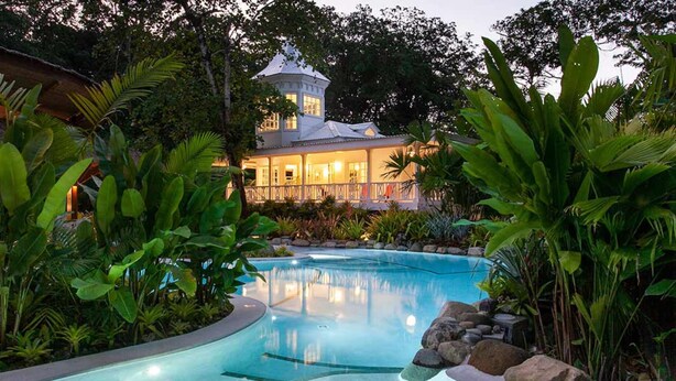 Una piscina rodeada de plantas tropicales, un edificio blanco con una torre y un jardín cuidado.