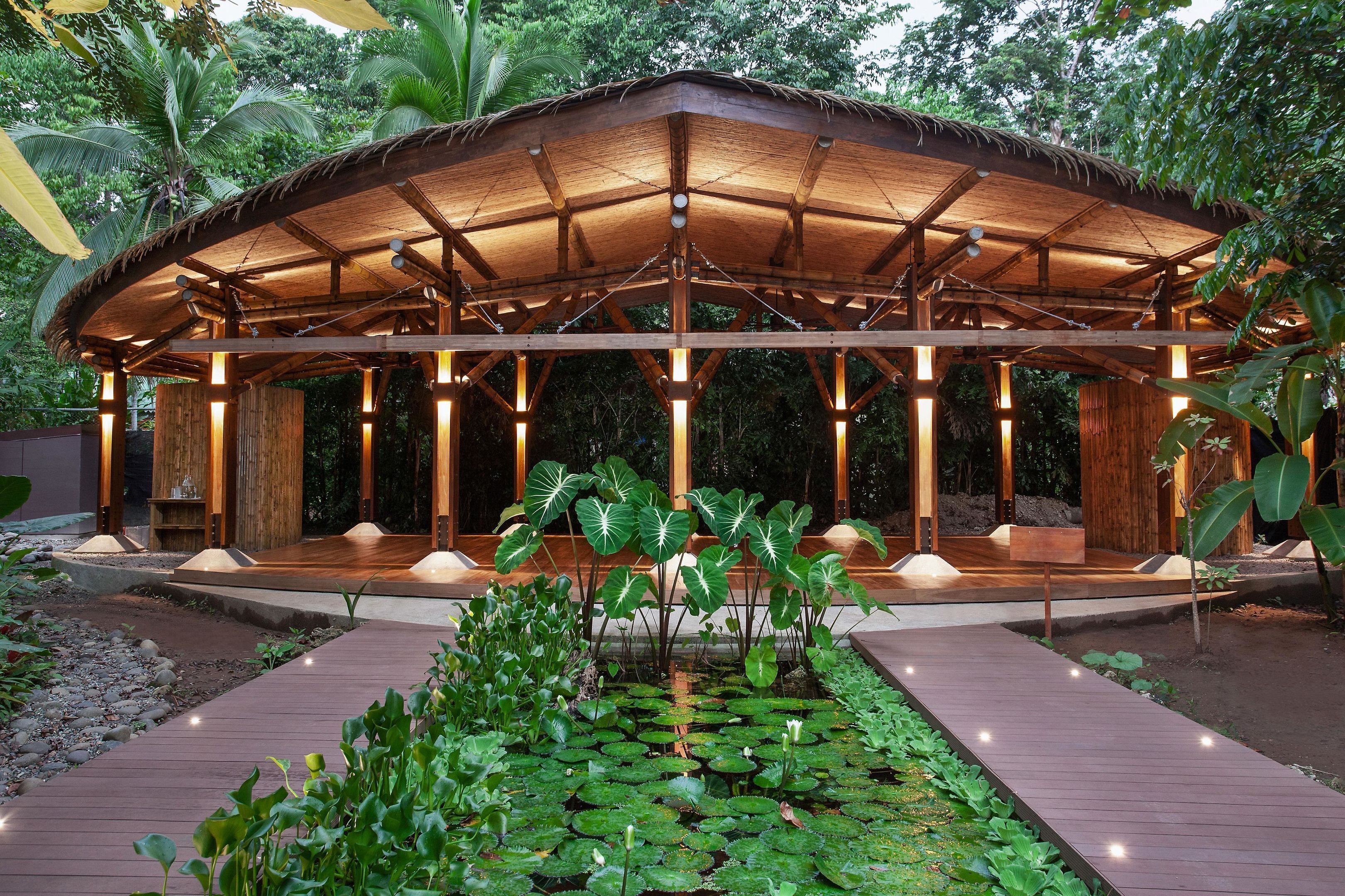 Un pavillon en bois, doté d’un toit de chaume, entouré d’un jardin où poussent des nénuphars et des plantes tropicales.