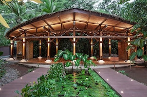 Un pabellón de madera con techo de paja, rodeado por un jardín con nenúfares y plantas tropicales.