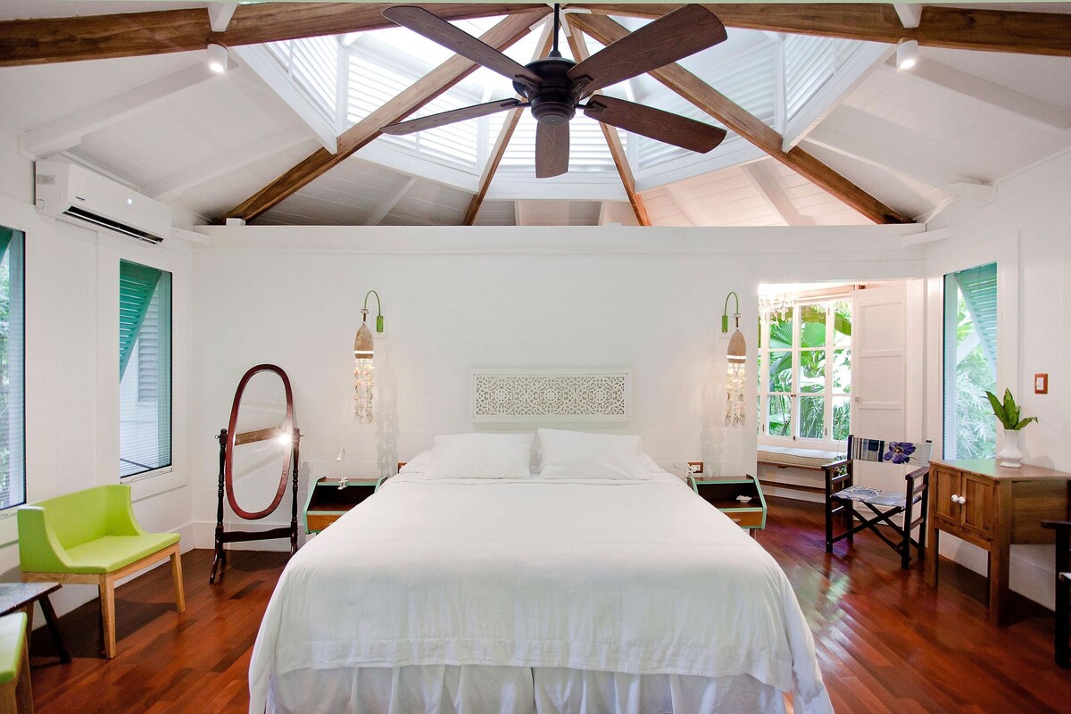 A bedroom with a large bed, wooden beams, a ceiling fan, and a skylight.
