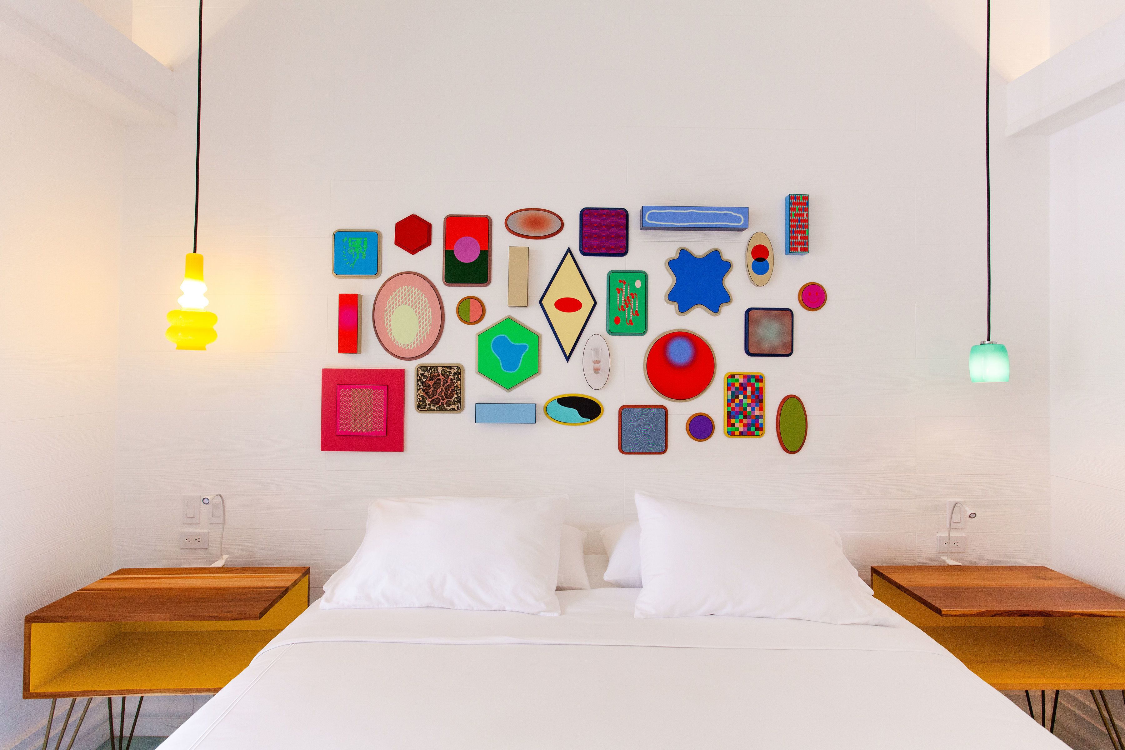Un dormitorio moderno con una cama, almohadas blancas y una pared decorada con formas geométricas coloridas. Hay dos mesitas de noche con tapas de madera y una lámpara colgante.