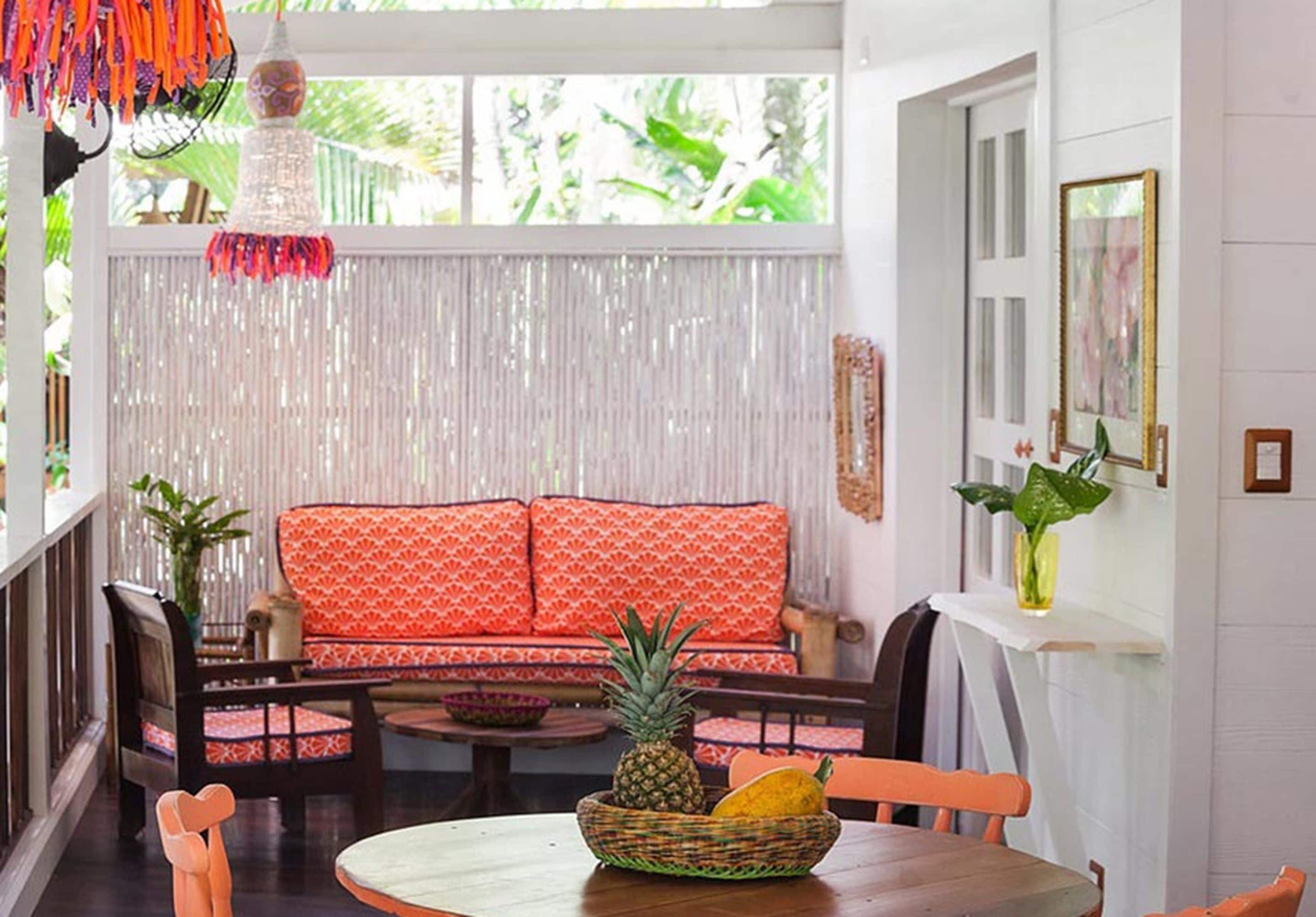 A room with a wooden table, a wicker basket with fruits, a red patterned sofa, and a white curtain backdrop.
