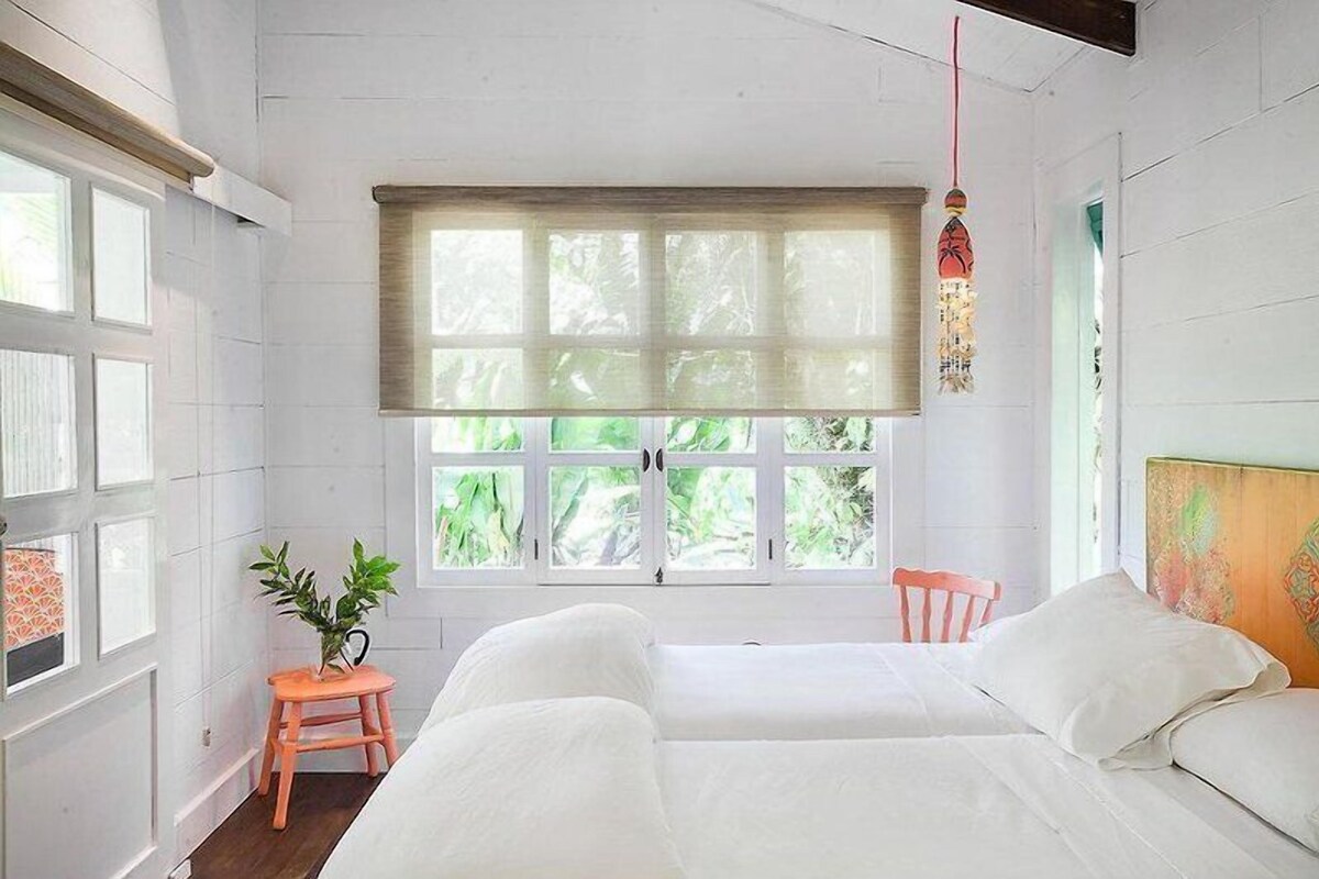 A room with a large window, a bed, a small stool, and a hanging decoration.