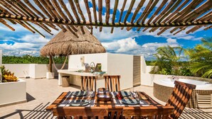 An outdoor dining area with a thatched-roof structure, a wooden table set for four, and a hot tub in the background.