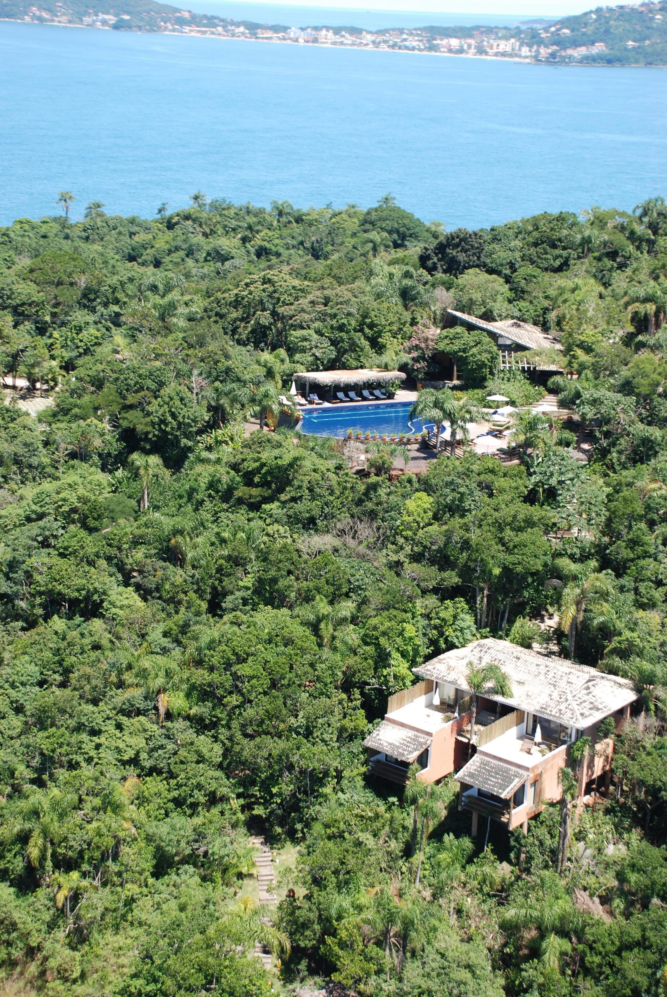 An aerial view of a resort with a swimming pool surrounded by dense greenery, a few buildings, and a view of the sea.