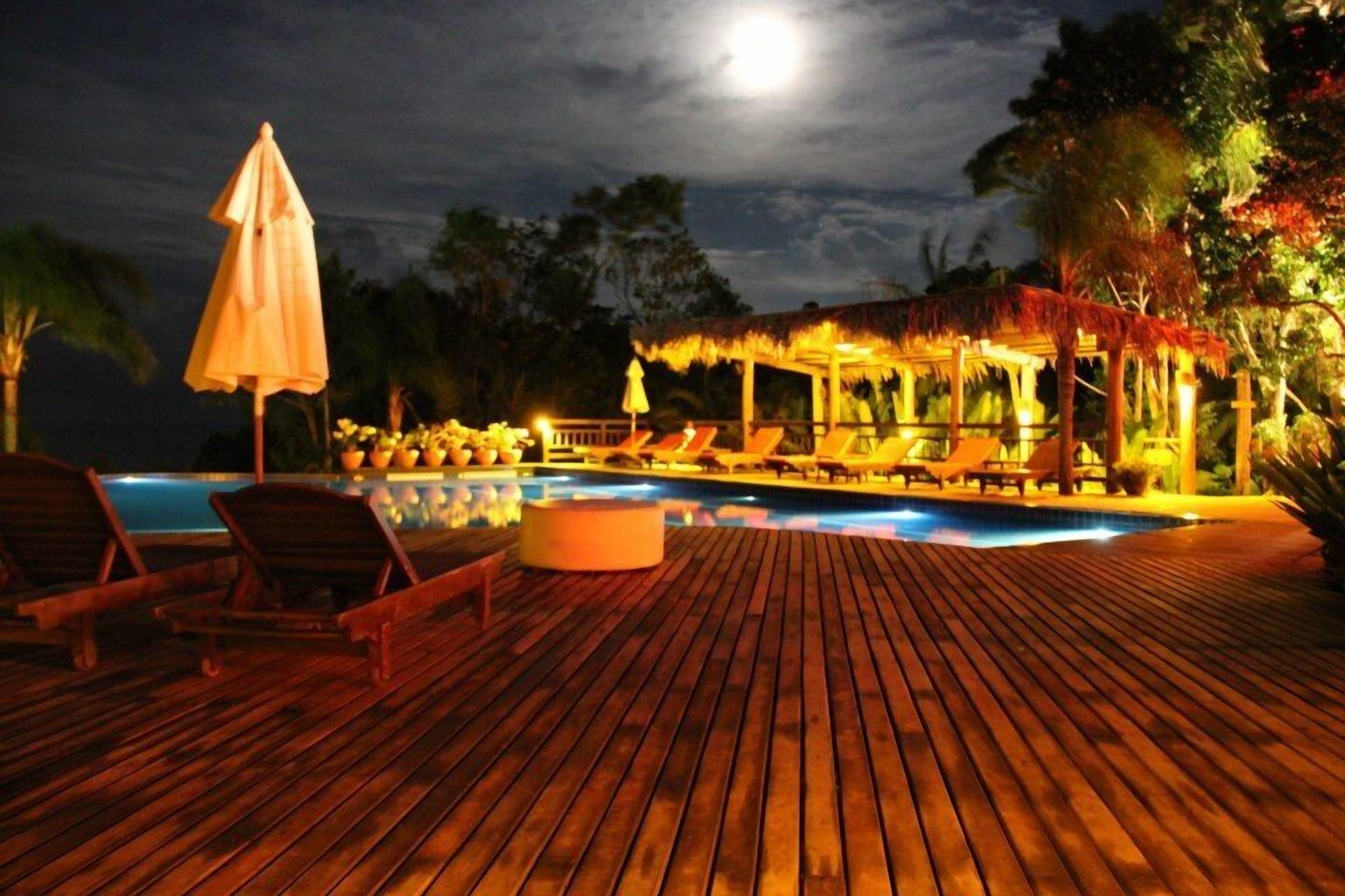 A wooden deck with lounge chairs and a pool area at night, illuminated by ambient lighting.