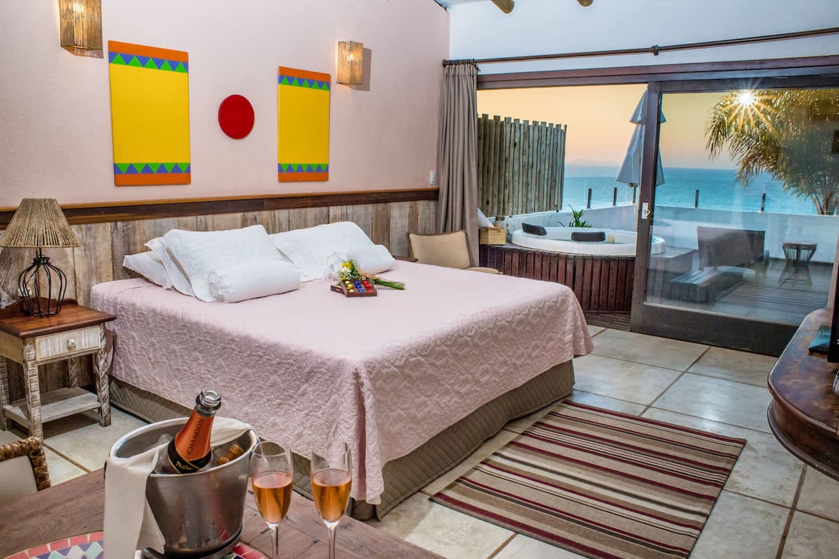 Una habitación de hotel con una cama grande, vistas al mar, un balcón con mesa y sillas, y una botella de champán sobre la mesita de noche.