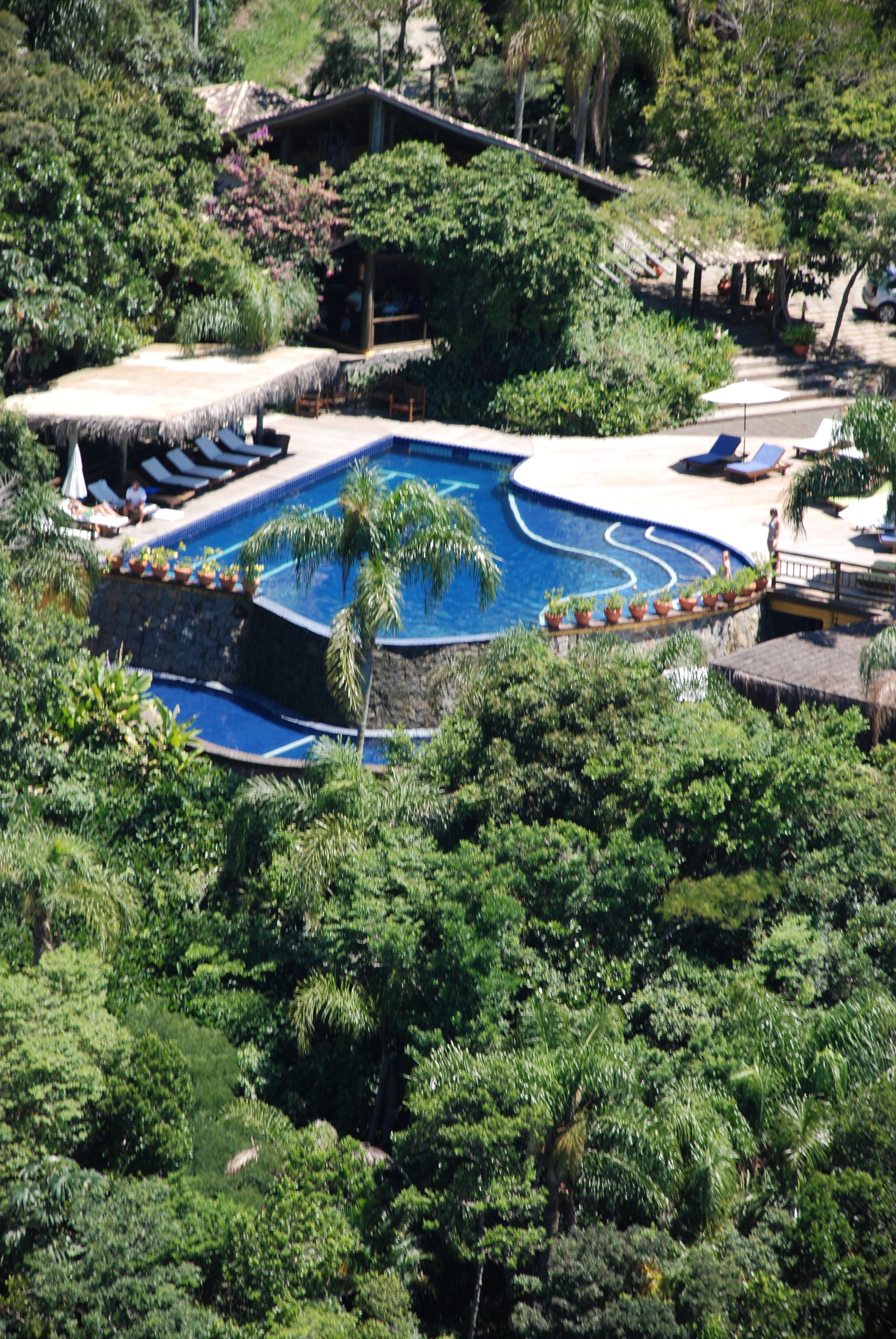 Ein Resort mit Swimmingpool, umgeben von üppiger Vegetation, einer Struktur mit Reetdach und Liegestühlen.