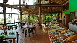 A spacious restaurant with wooden tables and chairs, a buffet line with various dishes, and large windows allowing natural light. - Refúgio do Estaleiro (Porto Belo)