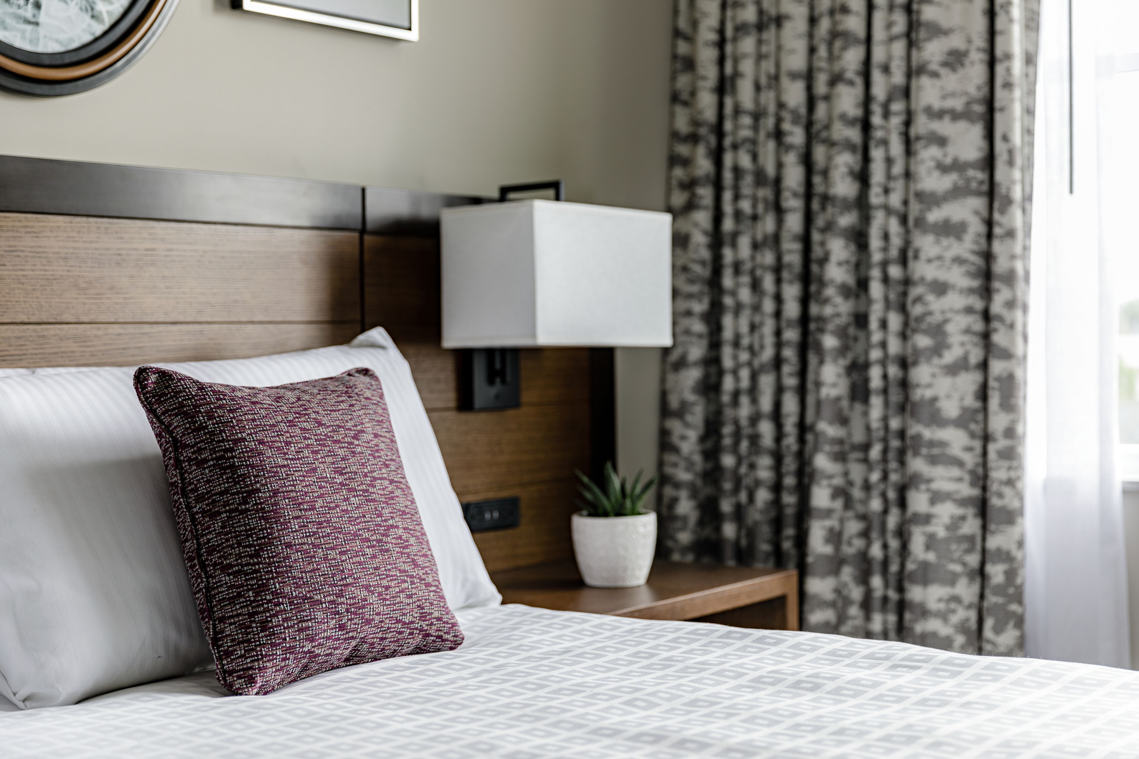 A neatly made bed with patterned pillows, a bedside lamp, a small potted plant, and a patterned curtain.