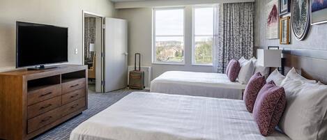 Habitación de hotel con dos camas, televisión, cómoda de madera y vista de la ciudad a través de amplias ventanas.