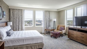 A hotel room with a large bed, a TV, a desk, and a chair. There are curtains on the windows, and a view of the city is visible.