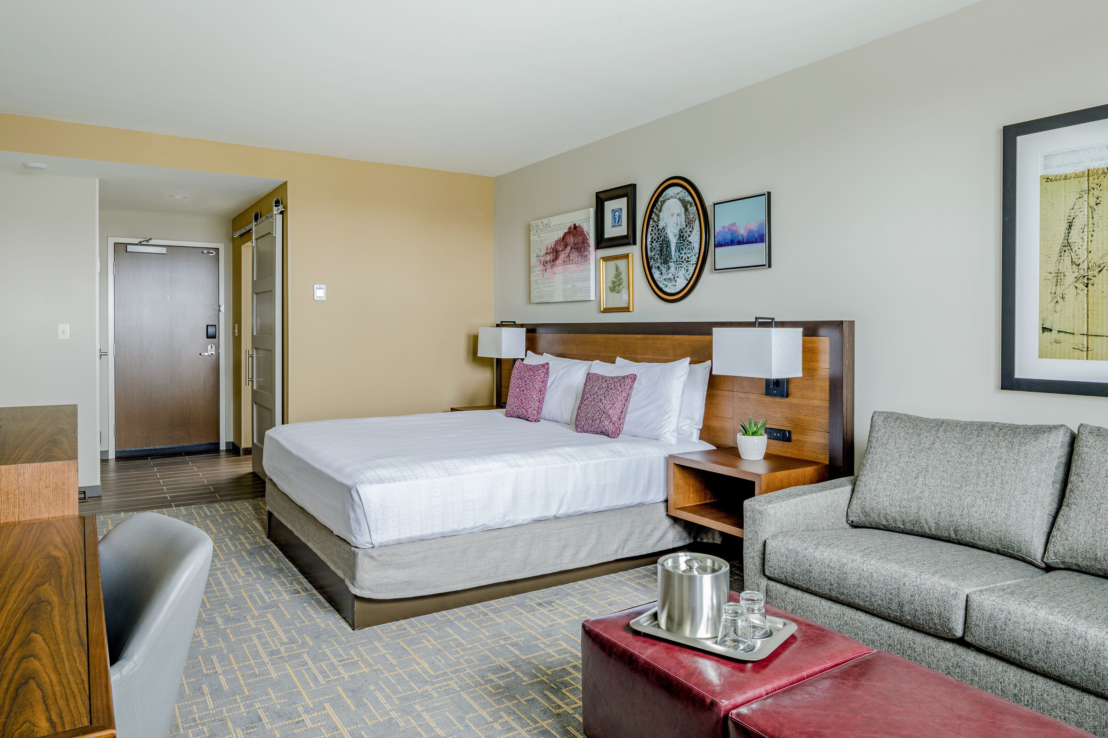 A hotel room with a bed, a sofa, a desk, and a coffee table. There are wall art pieces and a door leading to another room.