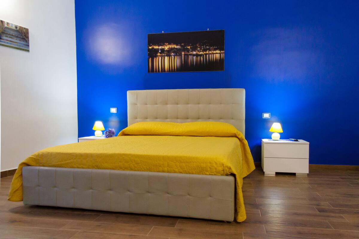 Een slaapkamer met een bed, twee bedlampen, een nachtkastje en een stadsgezichtschilderij aan de muur.