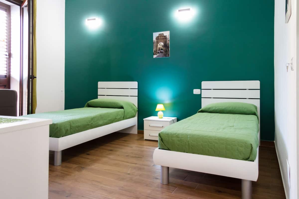 Een kamer met twee eenpersoonsbedden, een klein nachtkastje met een lamp, een aan de muur hangend schilderij en een raam met jaloezieën.