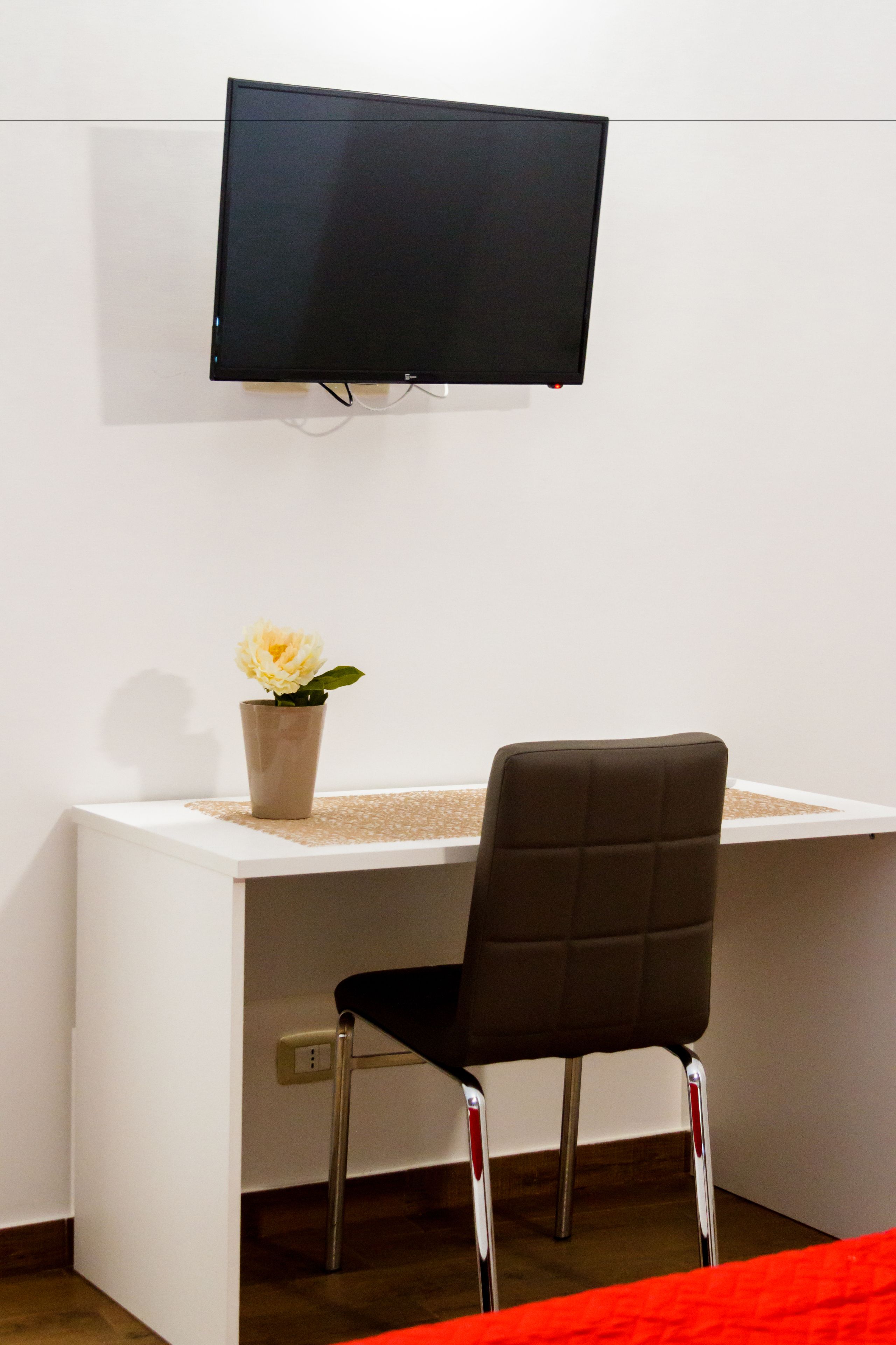 A room with a white desk, a black chair, a television mounted on the wall, and a vase with a flower on the desk.