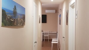 Un couloir avec un plancher en bois, des murs blancs et des portes. On y trouve un bureau avec des chaises et un téléviseur à écran plat fixé au mur.