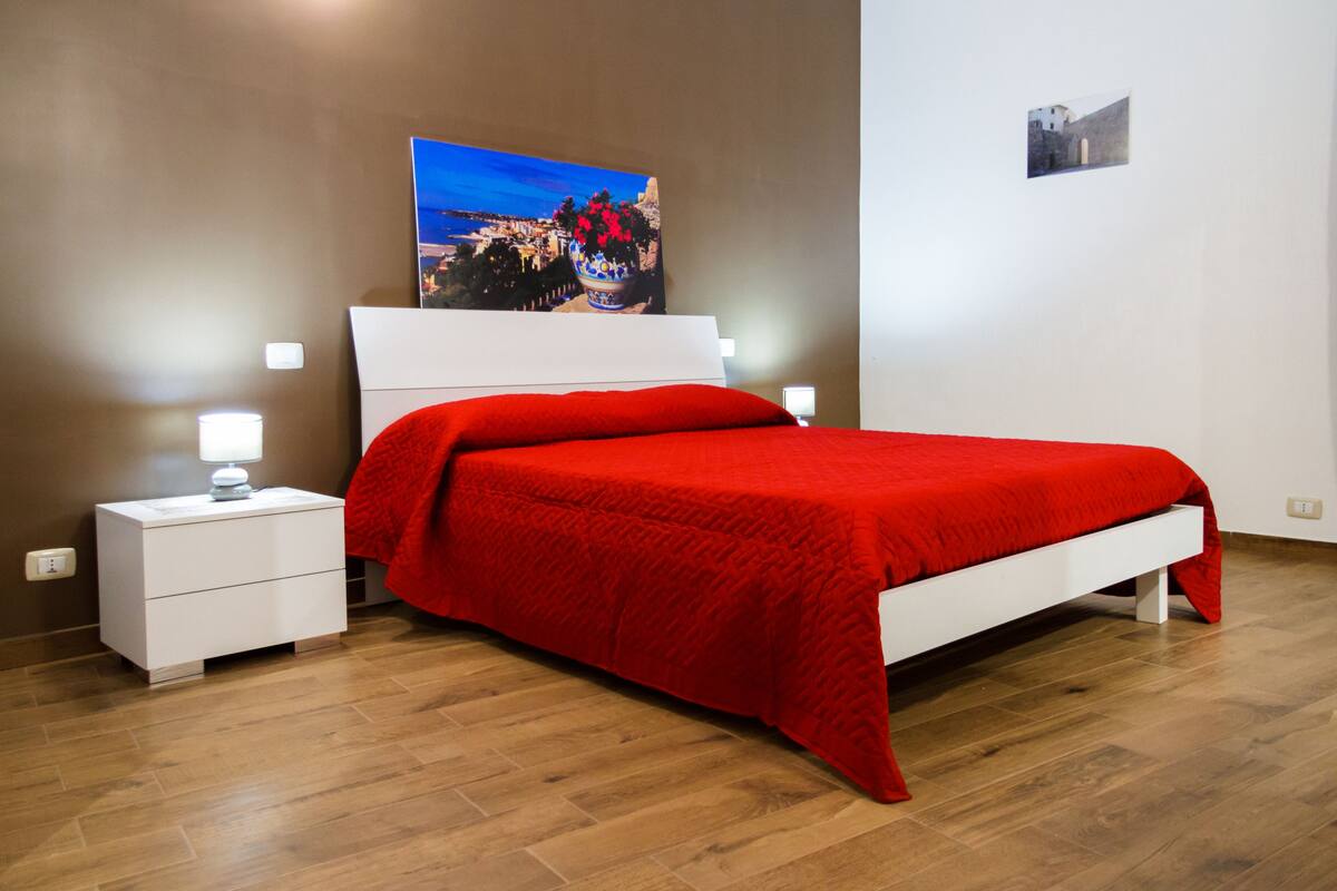 Een slaapkamer met een bed, witte nachtkastjes, een rode sprei, een schilderij aan de muur en een klein foto.