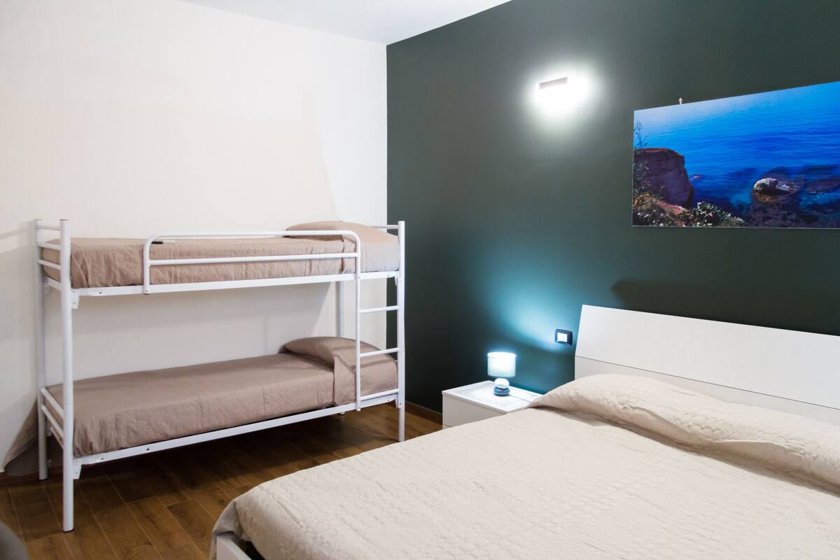Een kamer met een stapelbed, een enkel bed, een nachtkastje met lamp en een schilderij van een kustlandschap.
