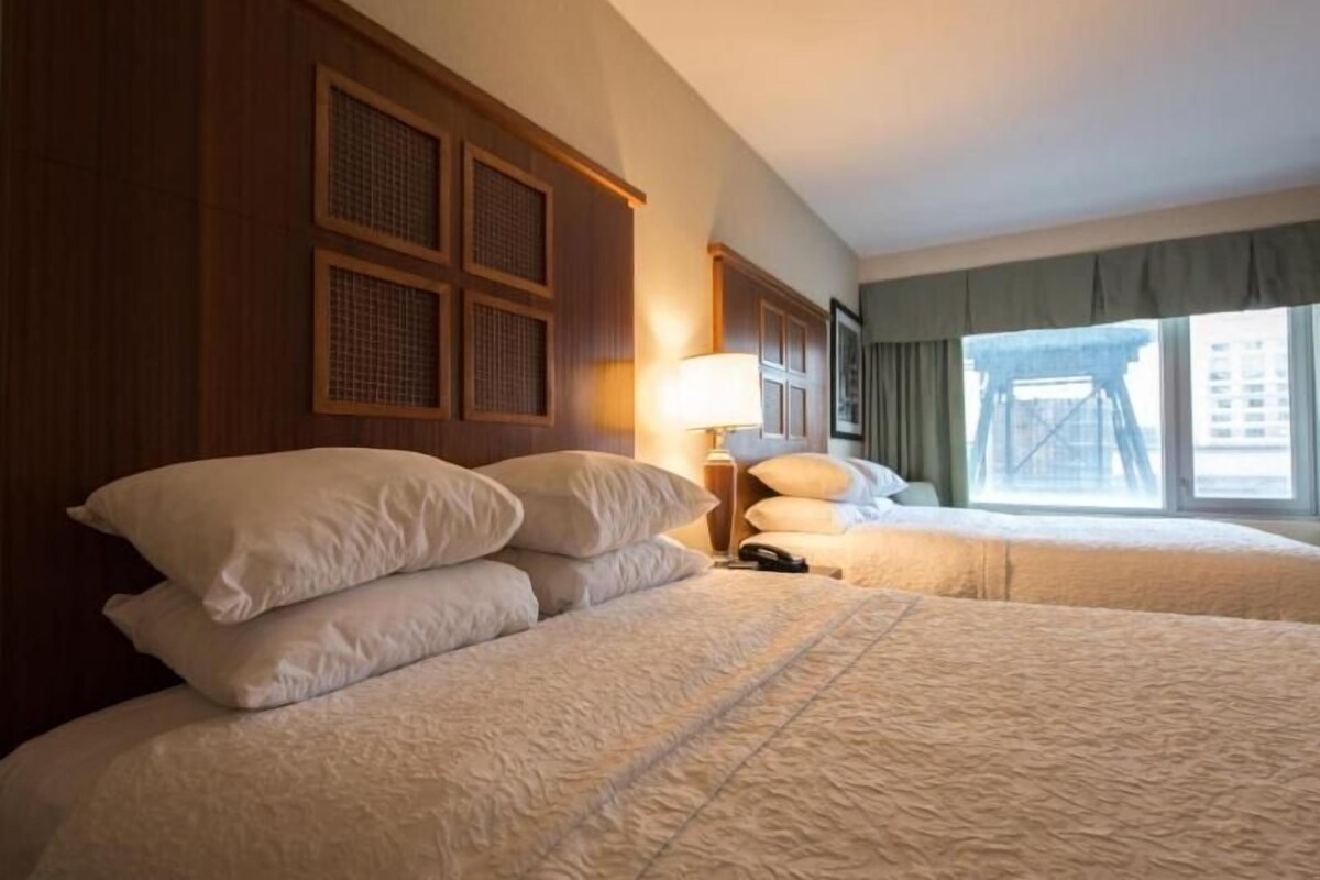 A hotel room with two beds, a wooden headboard, a lamp, and a view of a building outside.