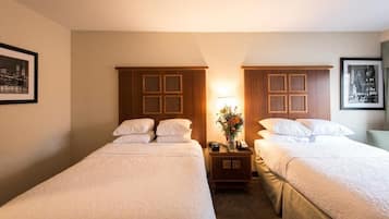 A hotel room with two single beds, a nightstand with a lamp and a vase of flowers, and framed pictures on the walls.