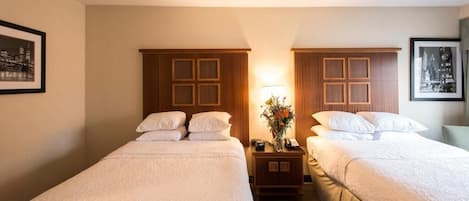 Una habitación de hotel con dos camas individuales, una mesita de noche con lámpara y un jarrón de flores, y cuadros en las paredes.