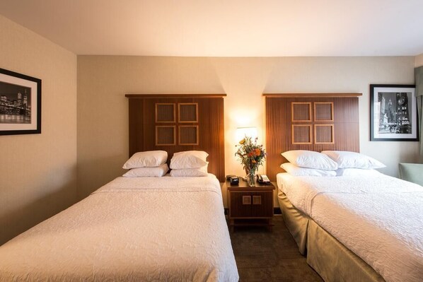 A hotel room with two single beds, a nightstand with a lamp and a vase of flowers, and framed pictures on the walls.