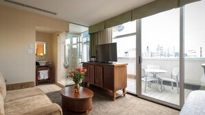 A hotel room with a sofa, a wooden coffee table with flowers, a TV on a wooden cabinet, and a balcony with a table and chairs.