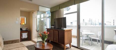 Een hotelkamer met een bank, een houten bijzettafeltje met bloemen, een televisie op een houten kast, en een balkon met een tafel en stoelen.