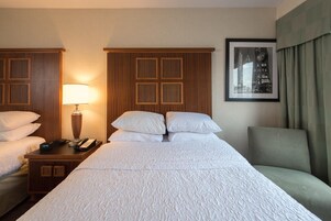 Quarto de hotel com uma cama grande, duas mesas de cabeceira, uma luminária, um quadro na parede e uma cadeira.
