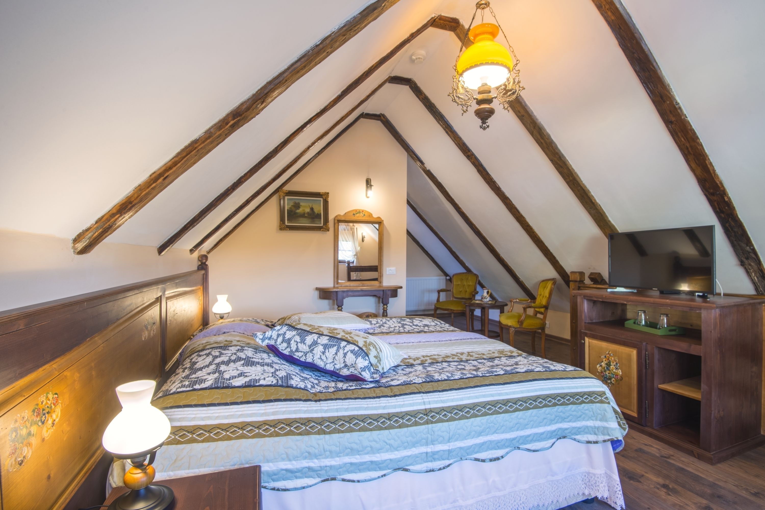 A bedroom with a sloped ceiling, a bed with patterned bedding, a bedside lamp, a chair, a television, and a wooden cabinet.