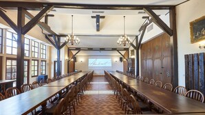 Een lange eettafel met stoelen, houten balken, kroonluchters en een projectiescherm in een ruime kamer.
