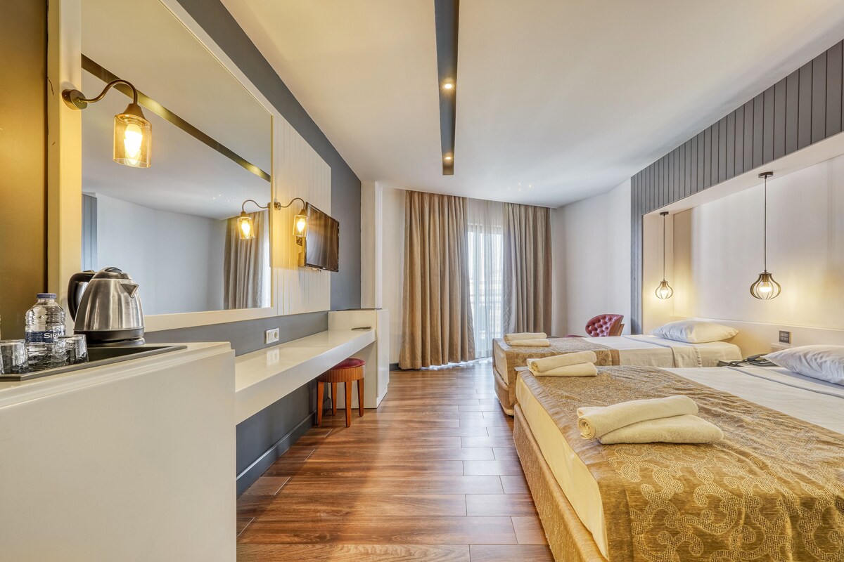 A hotel room with a large bed, a desk with a kettle, a chair, and a mirror.