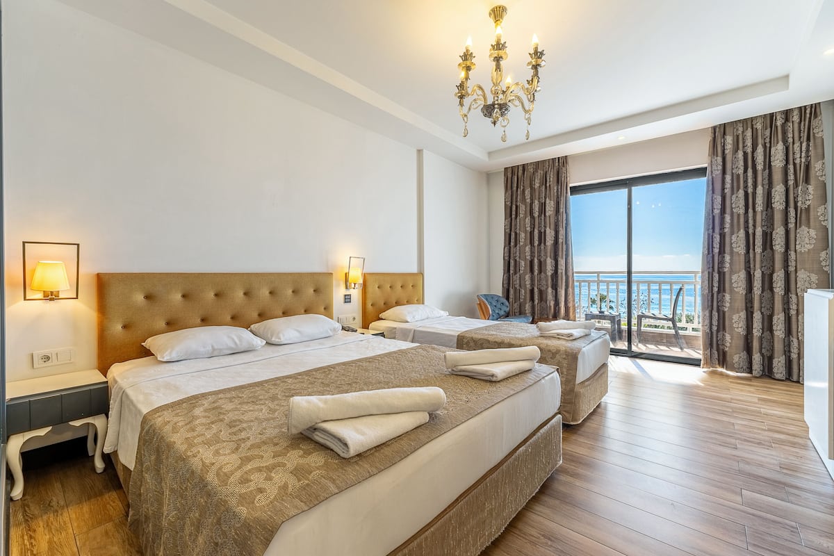 A hotel room with two beds, a chandelier, a balcony with curtains, and a view of the sea.