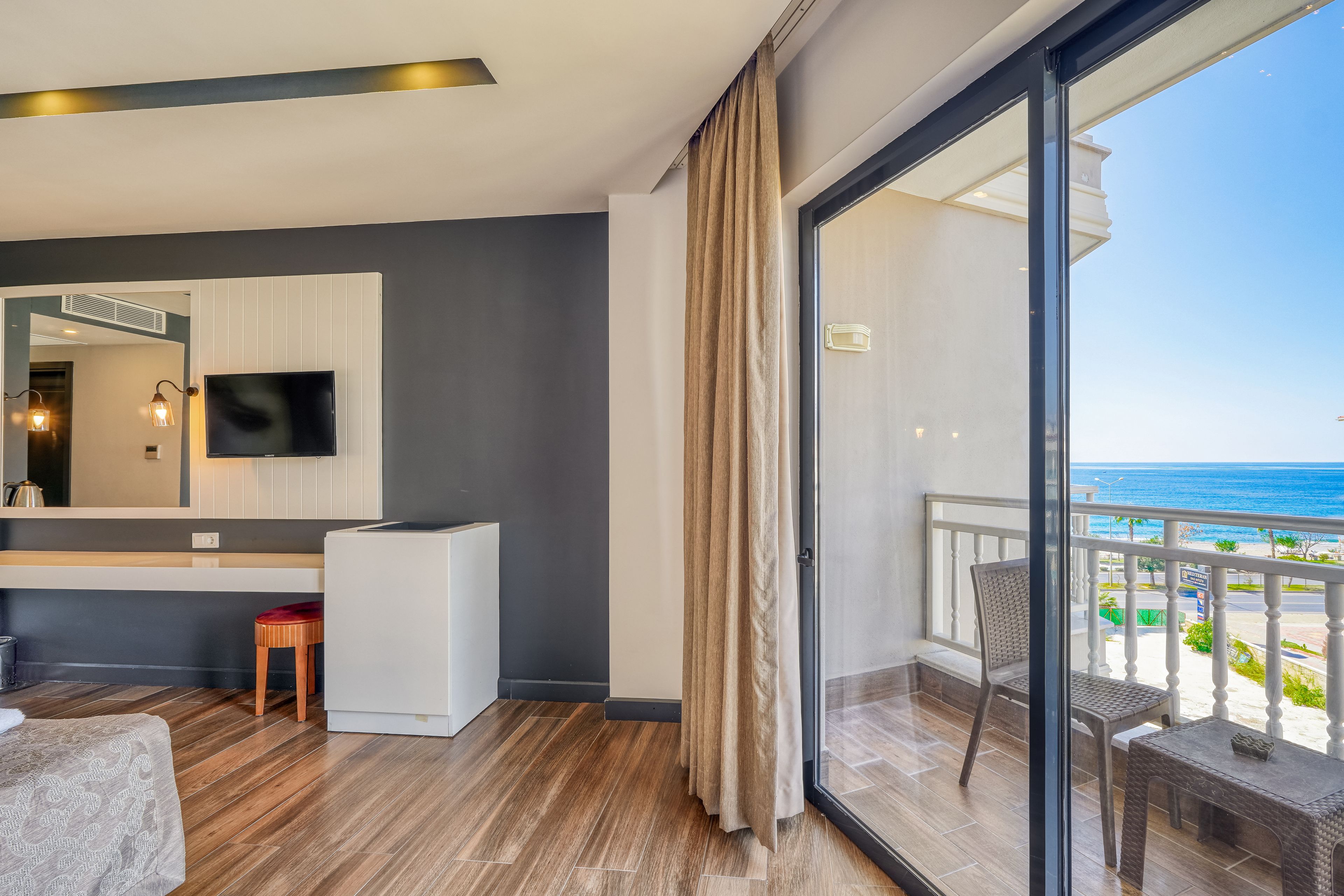 Una habitación de hotel moderna con un ventanal grande, un televisor de pantalla plana, un taburete de madera y un balcón con vista al océano.