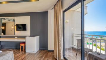 Una habitación de hotel moderna con un ventanal grande, un televisor de pantalla plana, un taburete de madera y un balcón con vista al océano.