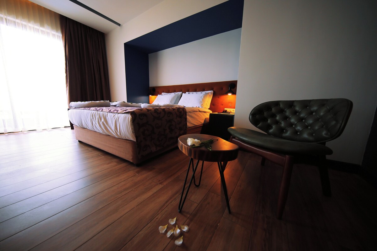 A hotel room with a large bed, a bedside table with a lamp, a chair, and wooden flooring.