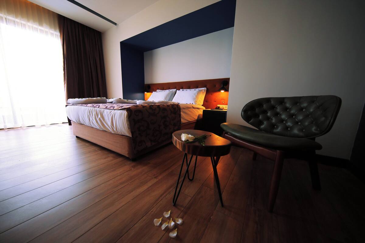 A hotel room with a large bed, a bedside table with a lamp, a chair, and wooden flooring.