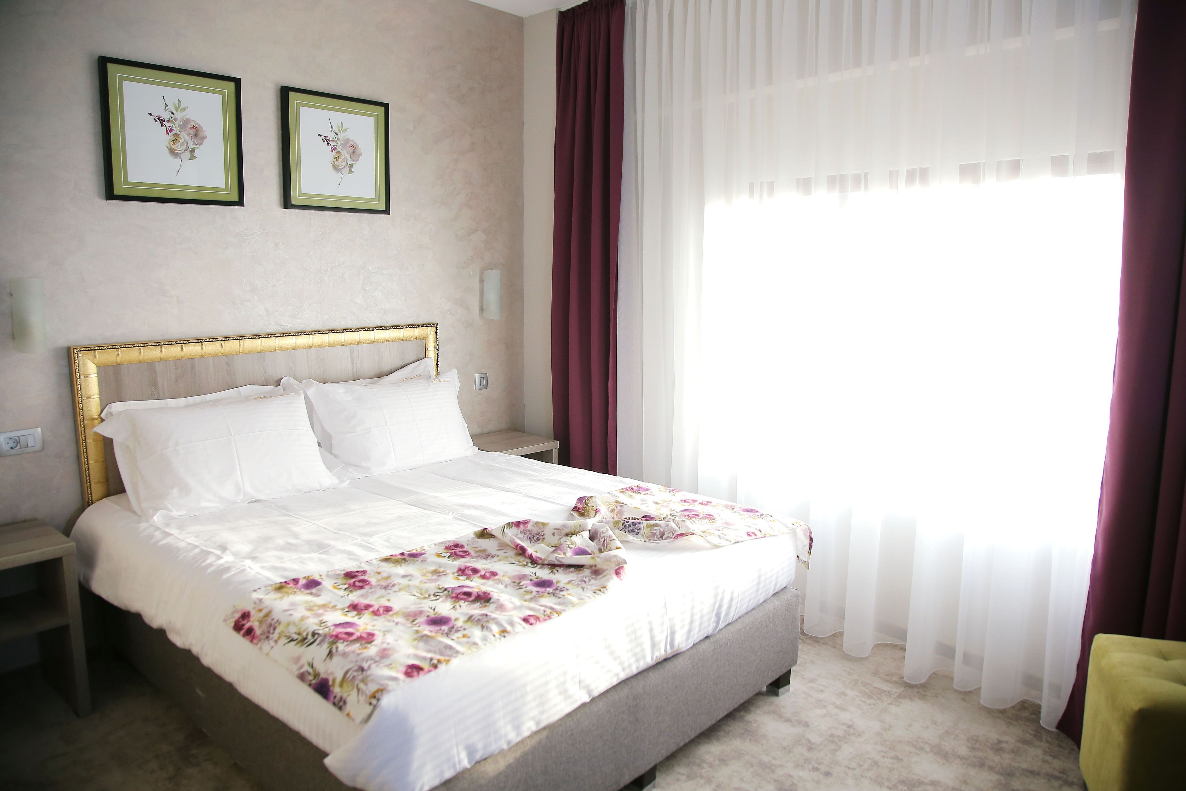 Una cama bien hecha con una colcha estampada con motivos florales, una mesita de noche y un revistero. Hay dos cuadros con flores enmarcados en la pared.