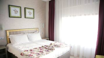 Una cama bien hecha con una colcha estampada con motivos florales, una mesita de noche y un revistero. Hay dos cuadros con flores enmarcados en la pared.